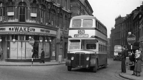 A black and white photo of an old number 50 bus on a street in Birmingham. People are wearing old fashioned, formal clothing and a woman is carrying a baby. There is another bus approaching in the background. You can faintly see passengers on the top deck of the main bus. 