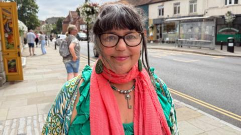 Amanda Gazidis standing on a pavement, smiling at the camera. A road and bus stop can be seen in the background, with a few people milling around. She is wearing a green top and pink scarf and has thick, dark-rimmed glasses on. 