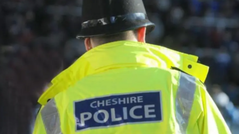 The back view of a Cheshire Police office. He wears a yellow hi-vis jacket with the force name in white letters on a blue background on the back and a standard police helmet.