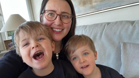 Kim Borthwick with her four-year-old twin sons Max and Freddie. She has long, dark hair and glasses and the boys have blond hair and blue eyes. They are sitting on a grey sofa in a living room and are all smiling at the camera 