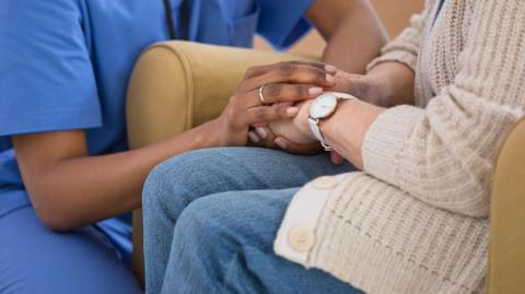 A nurse holding hands with an elderly woman. You cannot see either of their hands, the focus is on their hands.