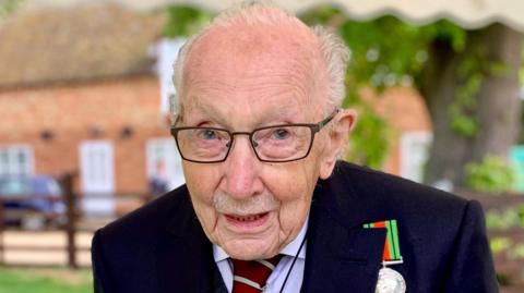 Colonel Tom Moore looks directly at the camera as he is photographed in a garden. He is wearing a dark jacket with a war medal attached on his left side. 