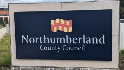 A large navy blue Northumberland County Council sign which has white lettering. Above the words is a Northumberland flag which has yellow and red squares. There is grass behind the sign and a brick building.