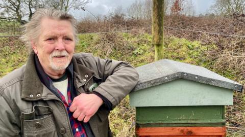 A man stands beside a homemade beehive in the middle of a field. The man has long grey hair and a thick green coat. He is resting his elbow on the hive. Behind the hive is a wire fence held up with wooden poles. In the distance is a large field with trees and shrubbery.