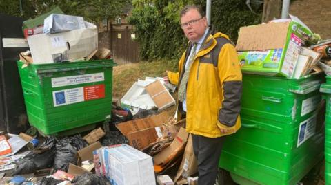 A man wearing spectacles, and a yellow waterproof coat over a grey suit,  stands in among piles of waste cardboard, in front of green recycling bins.