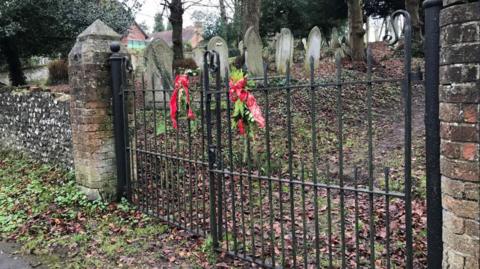 Two iron front gates mark the entrance to an overgrown graveyard. Several worn gravestones are pictured within the site which is overgrown with ivy and covered in fallen autumn leaves. There are brick walls either side of the gates. 