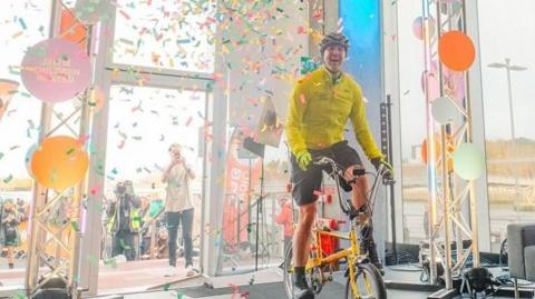 Paddy McGuiness in fluorescent cycling gear surrounded by confetti as he crosses the finish line on a kids' bike