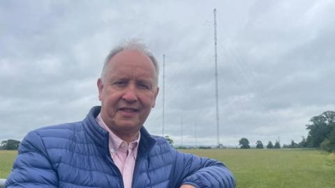 A man with grey hair, wearing a blue coat over a pink striped shirt stands in front of a green field with several trees. In the background you can see four tall transmitter masts with wires between them and coming down to the ground.