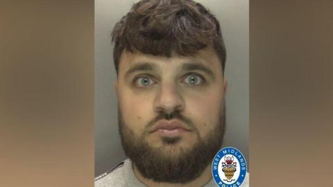 A mugshot of a man with a thick brown beard and light blue eyes. He has thick brown hair with a fringe and is staring into the camera. 