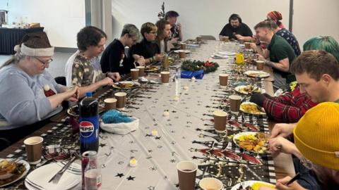 Thirteen people sitting at a long table eating Christmas dinner. There is a festive table cloth, brown paper cups, and plates full of food. There is a small Christmas tree behind one of the diners at the far end on the left.