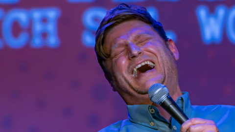 A man with blonde hair and a blue shirt holding a black microphone in front of a purple background with his head tipped back, laughing
