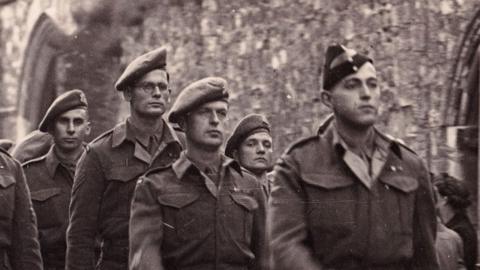 A black and white image showing men from the Cambridgeshire Regiment marching past Ely's war memorial in late 1945. They are in Army uniform. They are being watched by men, women and children.