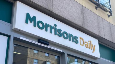 Photo of a Morrisons Daily store shop front in St Helier, Jersey. The sign is a white rectangle with blocked lettering reading "Morrisons Daily" in green and yellow.