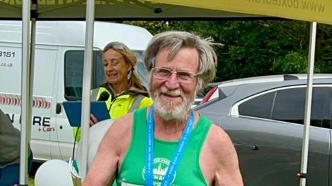 A joyful looking older man in a green running vest standing outside with cars parked behind him. He has a blue ribbon round his neck, is wearing glasses and has greying hair.