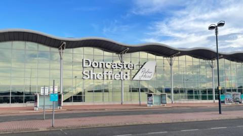 The terminal building of Doncaster Sheffield airport, A wide glass fronted building with a wavy roof is at the side of a road. There are two bus stops spaced out in front of the building. Large white letters on the front of the building read Doncaster Sheffield. A white sign to the right of them reads Robin Hood.