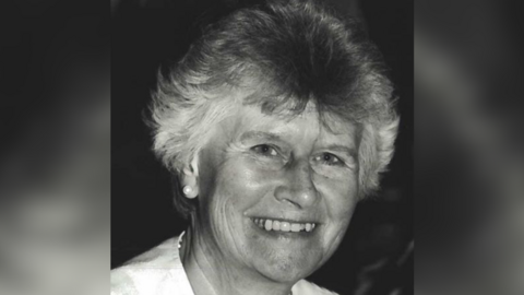 A black and white portrait photo of Beryl Purdy. She has short grey hair and is smiling at the camera. She is wearing a white top and pearl earrings.