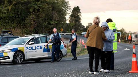 Two policemen standing near a police car, while two women wearing jackets and long pants stand on the side of the road. In the background is a tall green forest.