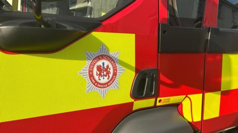 A fire truck is red and green in colour. There is a red and white logo which says Devon and Cornwall Fire Service.