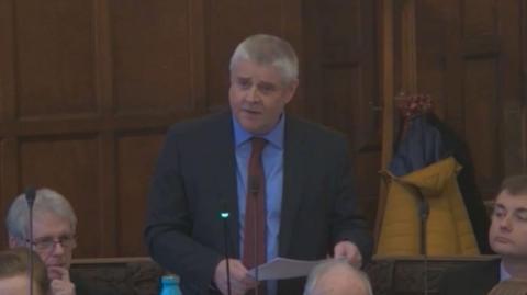 Peter Male stands in a wooden pannelled ornate council chamber speaking into a microphone. He wears a navy suit, blue shirt and red tie.