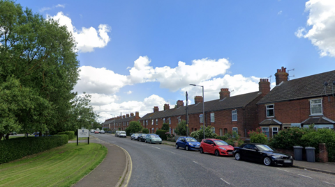 A residential street with a series of red-brick, two-storey houses with cars parked alongside the pavement at the front. On the other side of the road is a green patch of grass and trees. 