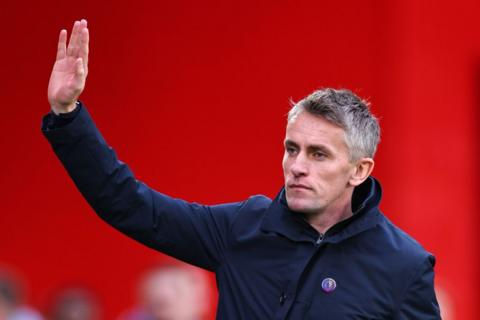 Kieran McKenna, with short grey hair, stands against a bright red background, looking to the side with a serious expression. He is wearing a dark navy blue buttoned jacket with a small circular badge on the left side of his chest, and his right hand is raised with his palm facing forward as if waving or gesturing.