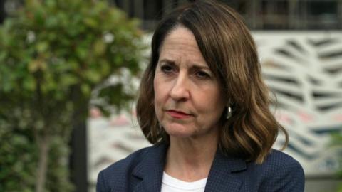 A medium close up of Pensions Secretary Liz Kendall wearing a blue jacket and white top with tree and bushes in background