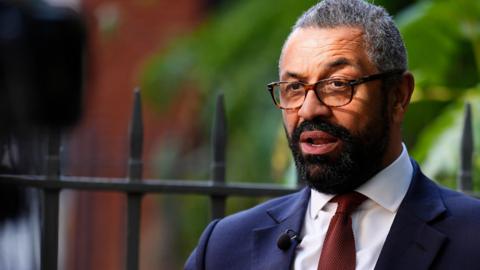James Cleverly giving a TV interview. He is standing in front of black railings with green vegetation behind him. He is wearing a dark blue jacket, white shirt and deep red tie. A white handkerchief in peeking out from his breast pocket. He has a neatly trimmed black beard, rectangular-framed glasses and short salt-and-pepper hair. He appears to be part way through answering a question, which he is delivering to a reporter and camera crew, which are just off frame to the left. He is wearing a small microphone on one lapel.