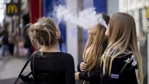 Three young women with long hair are standing in a street using vapes. They are exhaling plumes of vapour which looks like white smoke. 