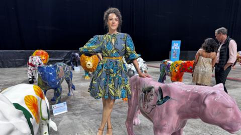 A woman, Natasha Clarke, stand among a display of animal sculptures, her hand rests on a pink lioness