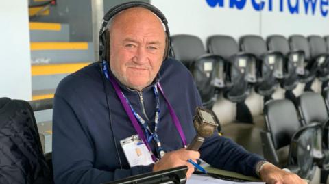 A man looks into the camera with a serious expression. He has is wearing headphones and around his neck is a navy blue lanyward with the BBC logo on it in white. In his right hand he is holding a radio microphone. Behind him are rows of stadium chairs in the upright position.