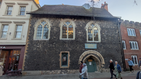 A very old building made of bacl bricks, which has three ornate windows and a very low doorway. People walk past on the street.