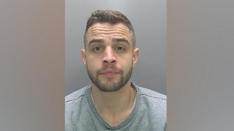 Nathan Giltinane is seen in a police custody photograph wearing a grey T-shirt. He is looking at the camera. He has brown eyes, short-cropped brown hair and a beard and moustache