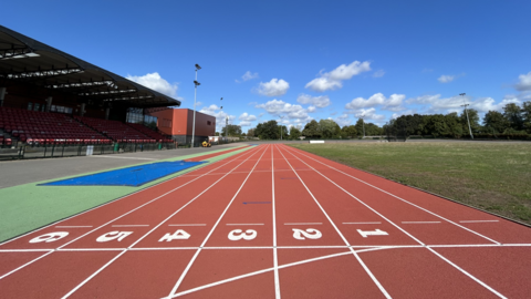 An orange, eight-lane athletics track underneath a blue sky. There's some raked seating on the left underneath a roof, and a stretch of grass to the right.