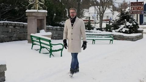 BBC Weather presenter Christopher Blanchett in a cream coat walking in the snow in Braemar, there are green benches behind him covered in snow