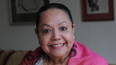 Candy Devine smiling at the camera as she sits on a sofa holding a guitar. She has her hair tied back and is wearing a pink shawl and a black and white patterned dress.