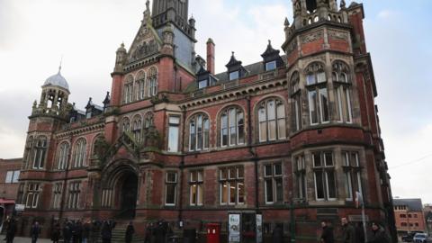 A grand Victorian redbrick building. A group of people are gathered around outside.