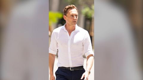 A man, Tom Hiddleston, in a white shirt and black trousers, looking wary.