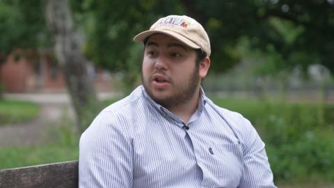 A man in a thin striped shirt and a cream baseball cap is sat on a public bench in a park. He has a thick but short beard. The background is a mixture of trees, grass and green plants with no flowers. 