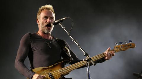 Sting holding a guitar and singing into a microphone on a stand.