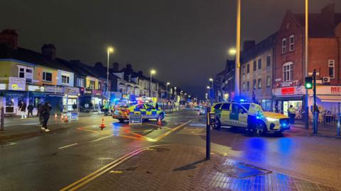 A high street lined with several shops is cordoned off by police. A police car and an ambulance car, both with flashing lights, are in the centre of the road.
