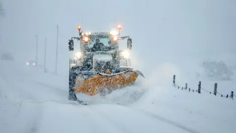 A snowplough drives through thick snow on the ground, as snow falls around it 