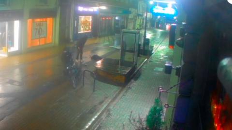 CCTV of Ryde High Street at night. In the centre of the frame you can see three figures. A woman lying on the ground a man standing over and a second man holding a bike. There is a phone box and a bench in the centre of the road. It appears to be raining.