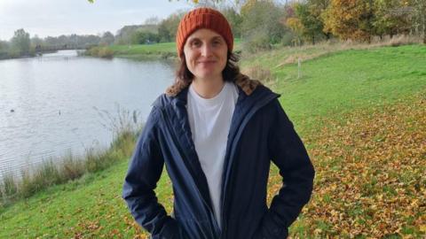 Rachel McGough stands in a park in front of a lake in an orange beanie and navy parker coat 