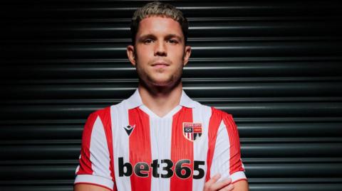 George Hirst poses with his arms crossed in Stoke's red-and-white striped home shirt