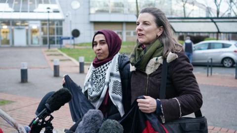 Two women standing in front of microphones outside court. The woman on the left is wearing a maroon headscard and Palestinian keffiyeh scarf around her neck. The woman on the right has shoulder length brown hair and is in a dark coat. They are about to hold up a dark banner.