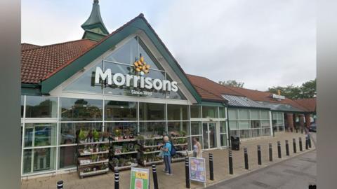 The front entrance of Morrisons, with a large sign on the triangle shaped front.
It has a red tiled roof and green edging, and glass front. Stands of plants are outside the front, which has a paved area lined with bollards. Two people stand looking at the plants.