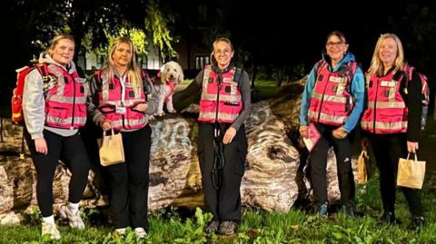 Five women in bright pink high-vis jackets stand smiling on night patrol, with small white dog Blossom in a matching vest.