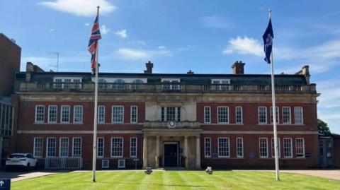 The front facade of Wootton Hall police headquarters. It is a three storey building set in front of a neatly presented lawn with two flag poles. 