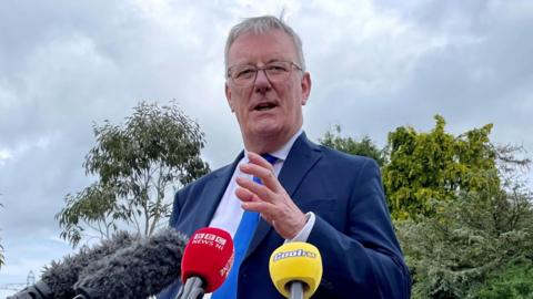 Mike Nesbitt speaking into media microphones. He is shot slightly from below so the sky is behind him and he is wearing a blue suit.