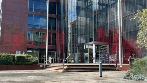 Large mainly glass-fronted office building with steps up to a glass-fronted entrance.  The wing to the left is coloured red.  There is red paint on several windows up as far as the second floor.  There are bushes in the foreground on either side. There is yellow tape across the entrance, and doors appear to have been smashed.